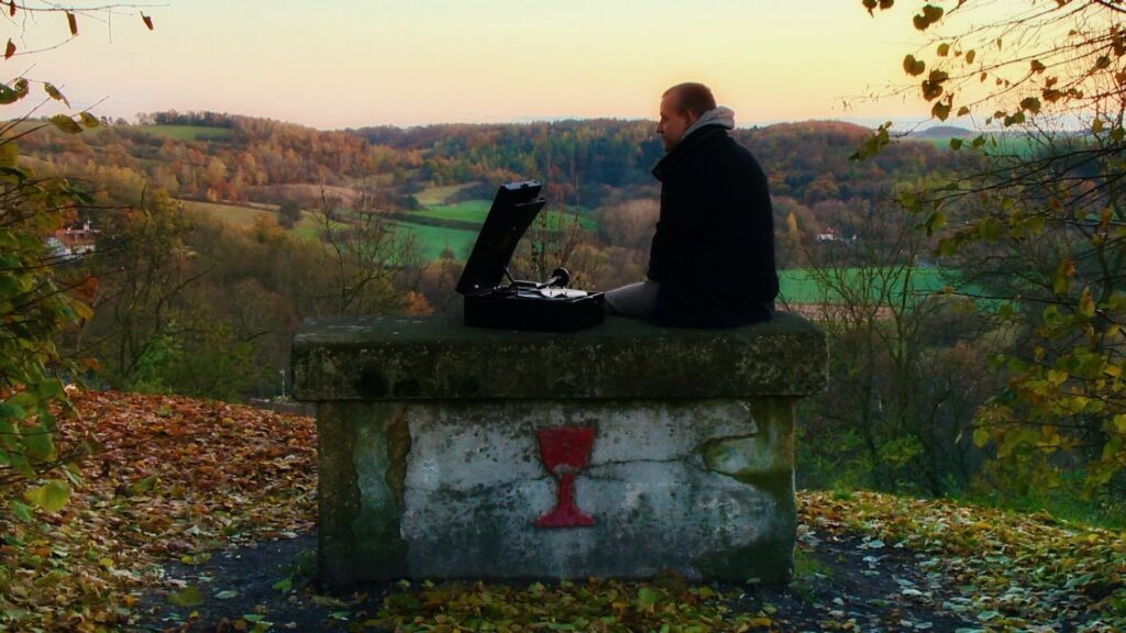 Muž sedí s gramofonem na betonovém stole nad podzimní krajinou — záběr z filmu Tekno není všekno
